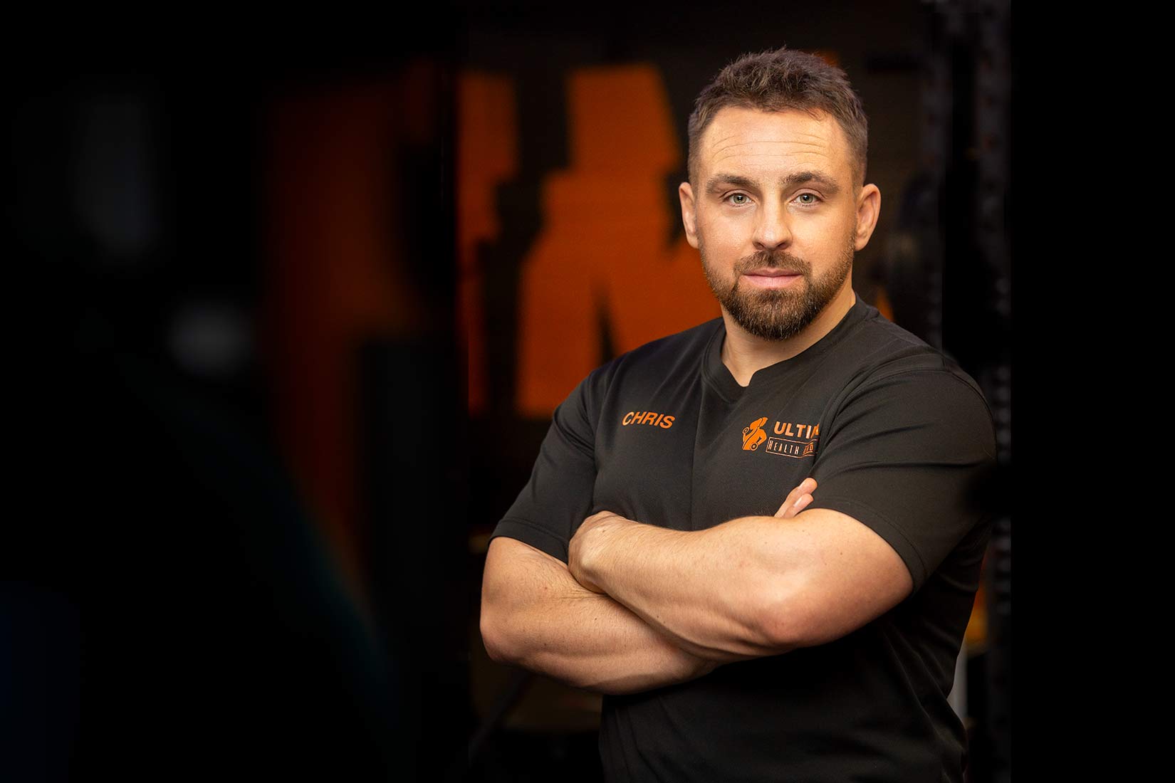Personal Branding style portrait of a gym owner and coach wearing a black t shirt with arms folded against a dark background. Also called and environmental portraits style