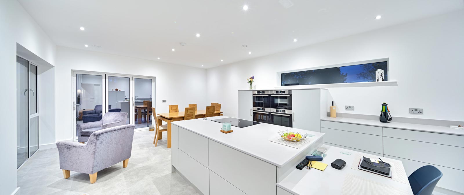 Stark white kitchen interior with large island and armchairs looking towards bifold doors by interior photographer Matthew Jones