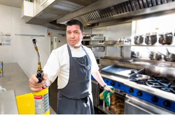 Business portrait of chef with blue apron holding a blow tourch with a kitchen background
