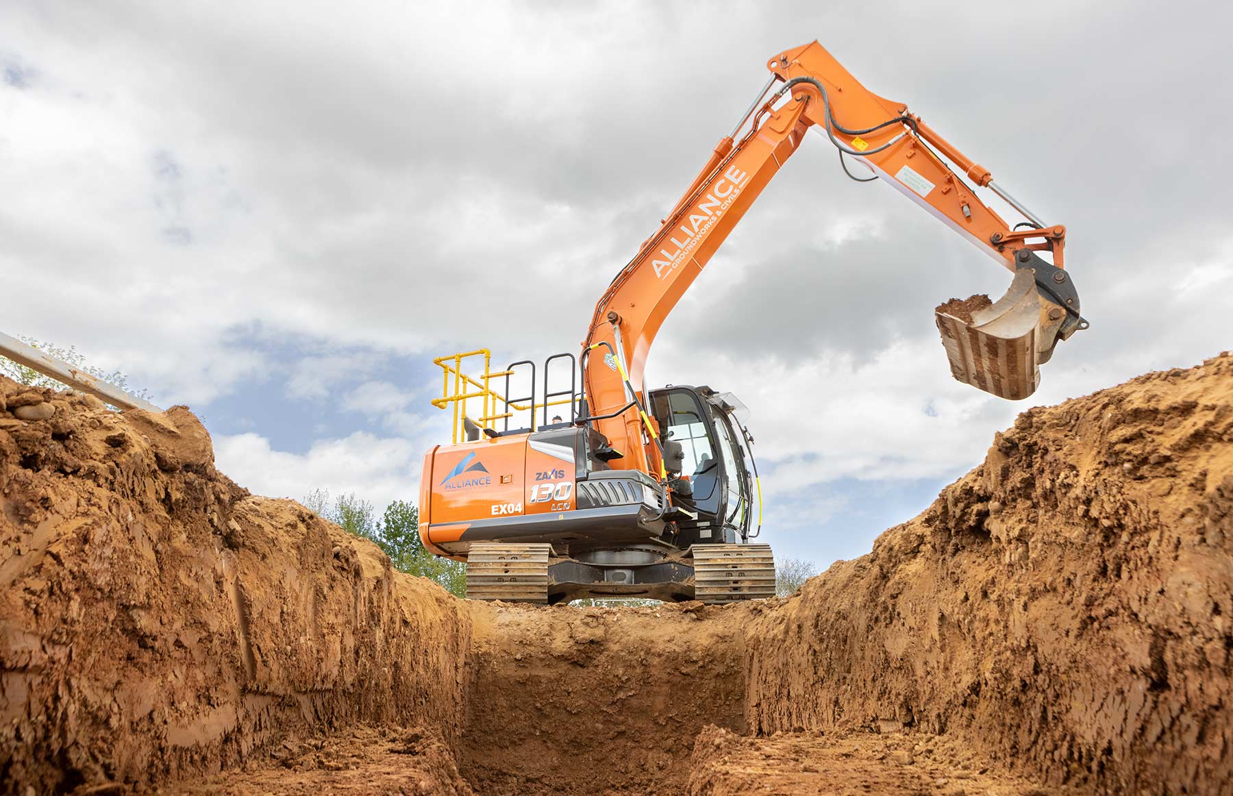 An orange Zaxis 130 excavating a trench against a cloudy sky