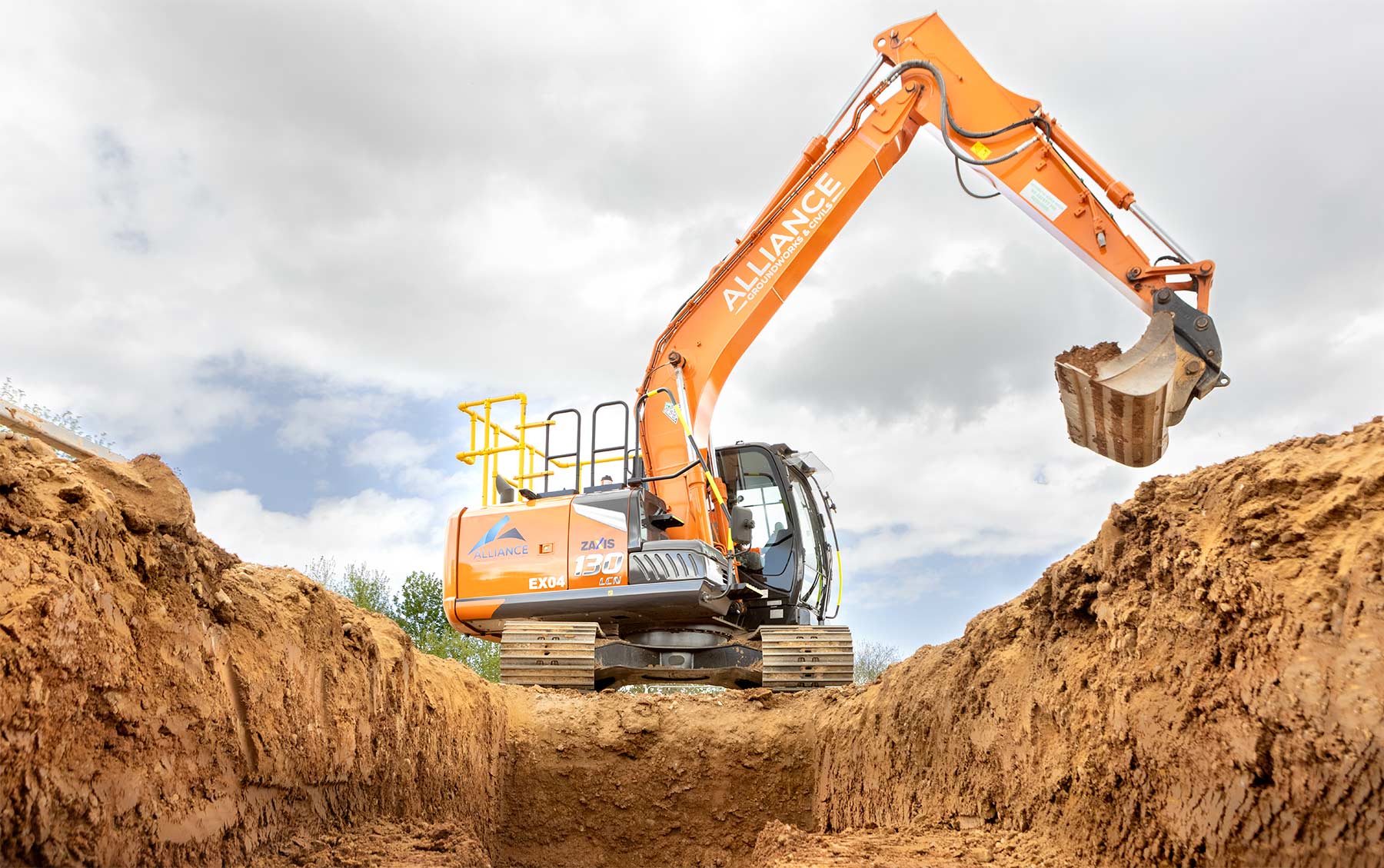 An orange zaxis 130 against a cloudy sky digging a drainage trench in brown earth