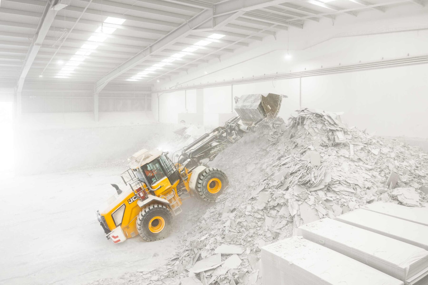 A front loader a full stretch pushes plasterboard in an industrial recycling facility to the top of a pile. It's front wheels are half way up the slope.