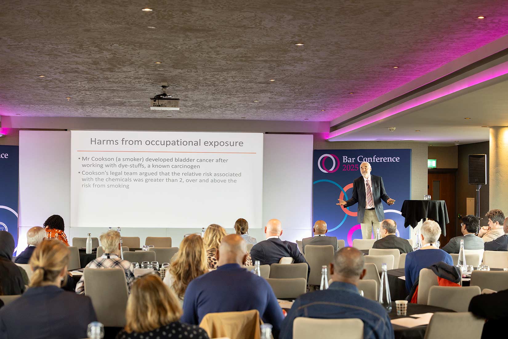 Full length keynote speaker on stage at a Manchester conference, presentation slide and event branding clearly visible behind.